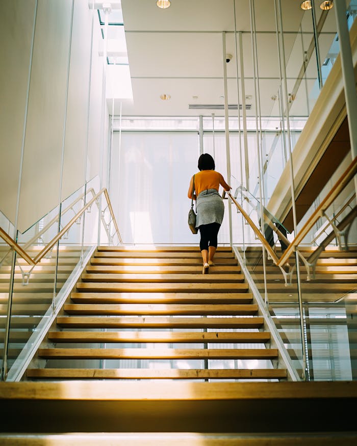 A woman walks up a well-lit modern staircase with glass railings inside a building.